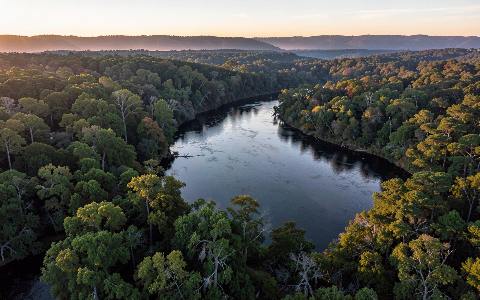 Aerial view of a wide slow river curving through dense hardwood forest at sunrise